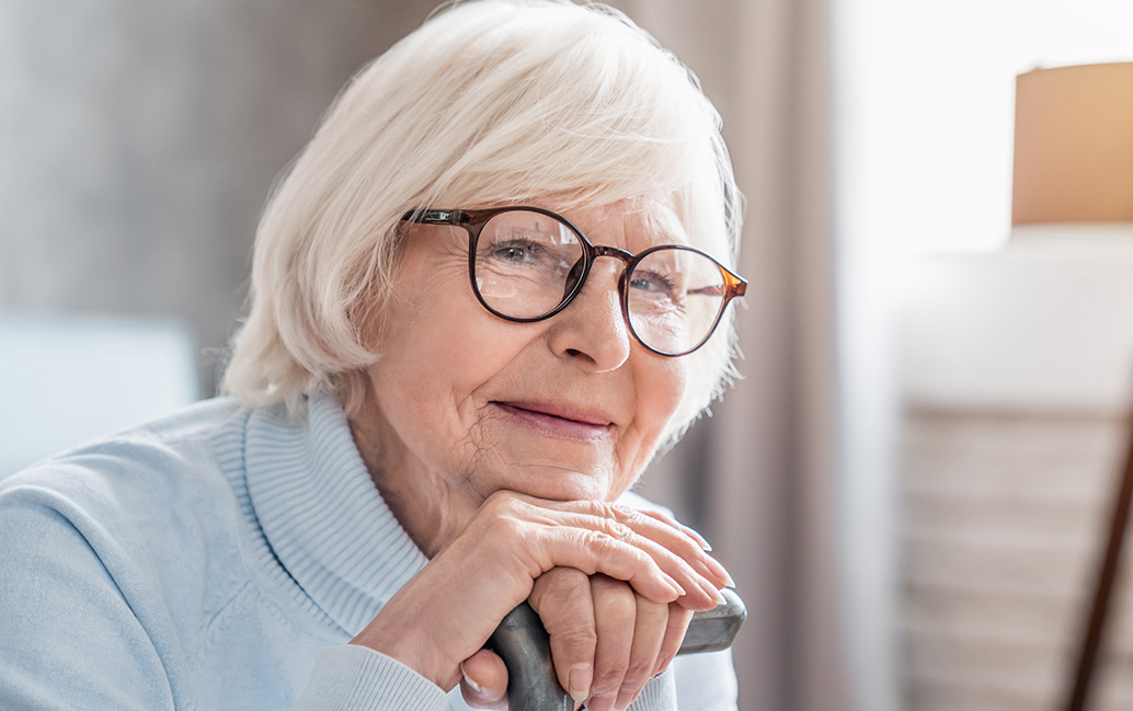 Elderly lady wearing glasses and smiling while leaning on her cane